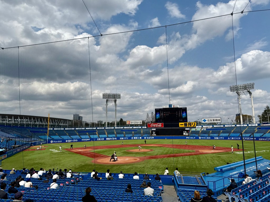 A spacious baseball stadium viewed from the stands, featuring a game in progress on the field, surrounded by empty blue seats and cloudy skies.