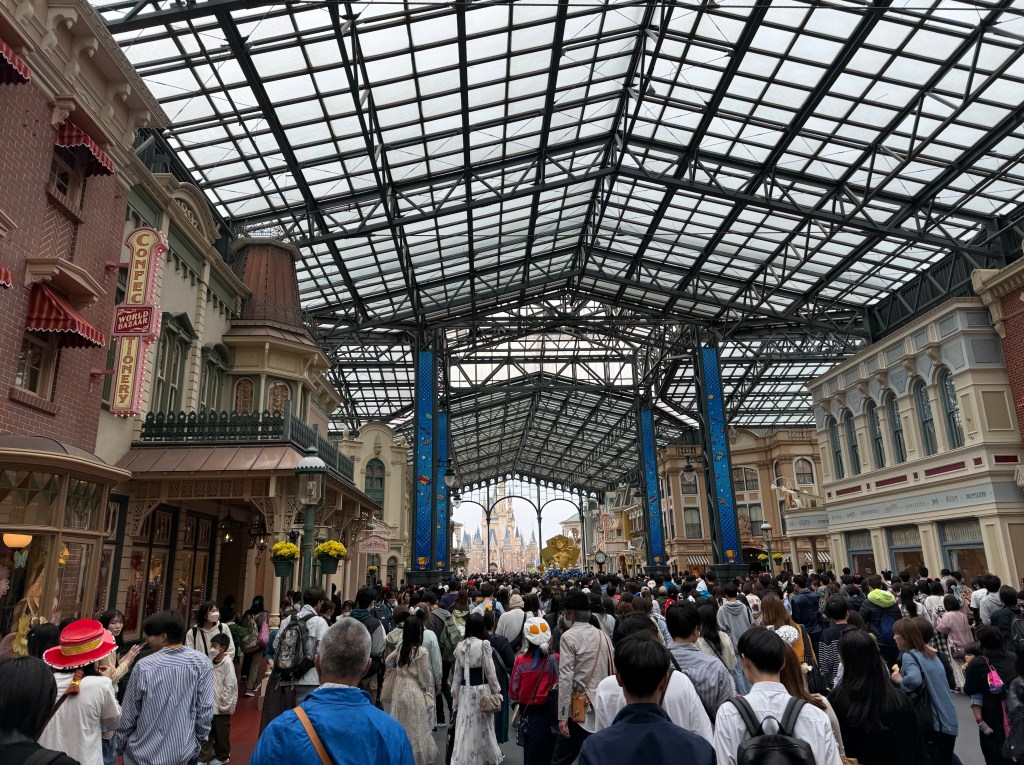 Crowd of visitors under a large glass-covered area in Tokyo Disneyland, showcasing themed buildings and decorations.